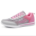 A women's athletic sneaker in gray and pink, featuring a perforated mesh upper, pink laces, a pink heel counter, and a white sole.