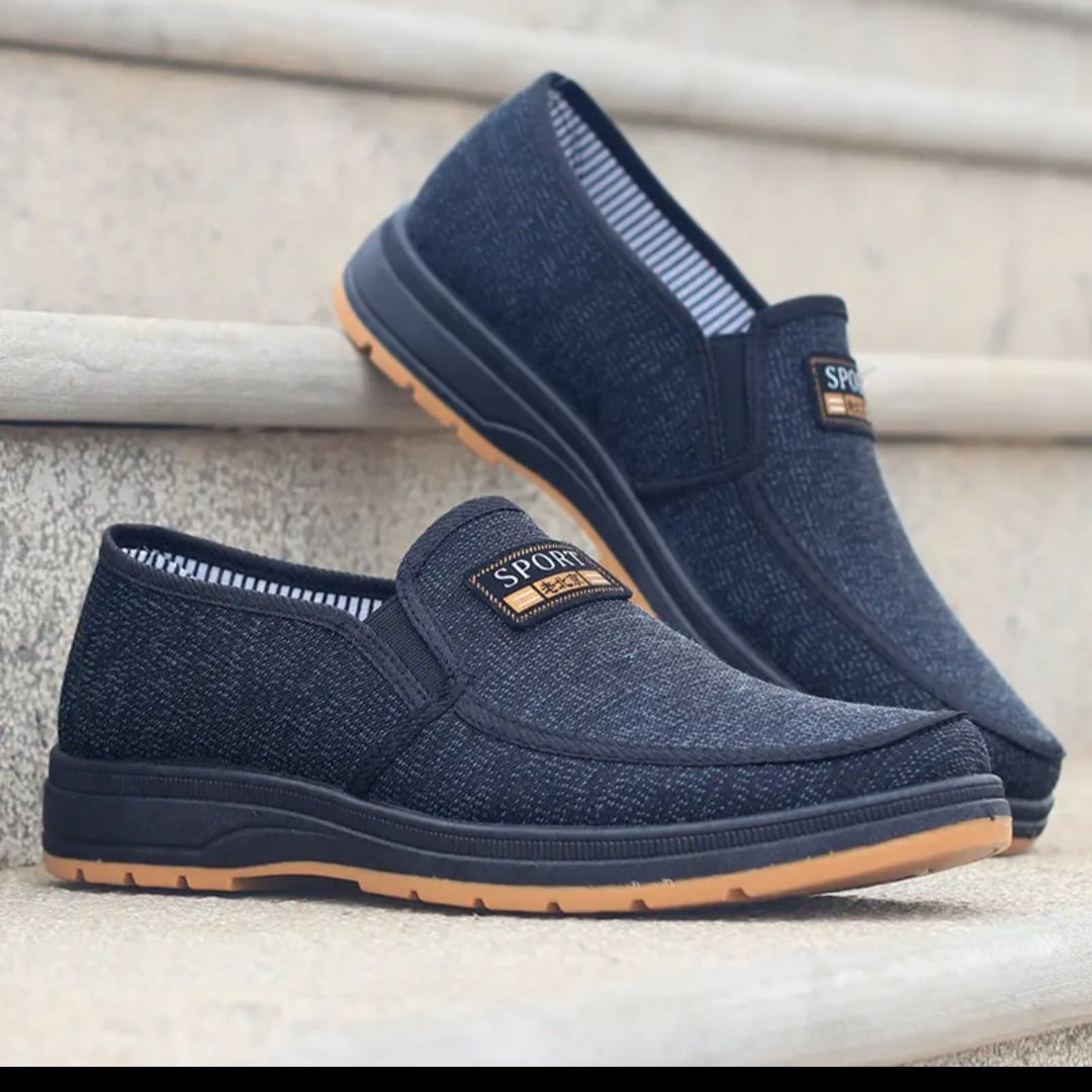 A pair of dark navy slip-on casual shoes with a textured fabric upper and a tan outsole, displayed on stone steps.