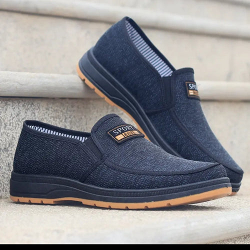 A pair of dark navy slip-on casual shoes with a textured fabric upper and a tan outsole, displayed on stone steps.