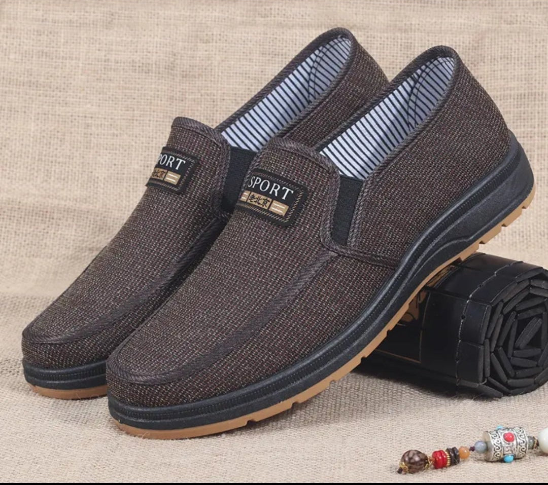 A pair of men's casual slip-on shoes in dark brown textured fabric with a striped blue and white lining, displayed on a beige textured background with a rolled black rug prop.