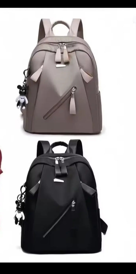 Two backpacks shown on a plain white background: one taupe-gray and one black, each with top handles, front zip compartments, side pockets, and hanging keychains.