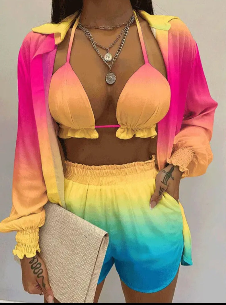 A woman models a two-piece swimwear set with a matching long-sleeve open shirt. The set features a gradient color scheme transitioning from pink and orange on the top to yellow, green, and blue on the shorts.