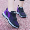 A pair of women's athletic sneakers in black and purple, shown on a person's feet on a pinkish-red textured surface. The shoes feature a black mesh upper with purple side panels, black laces, and a black and purple sole.