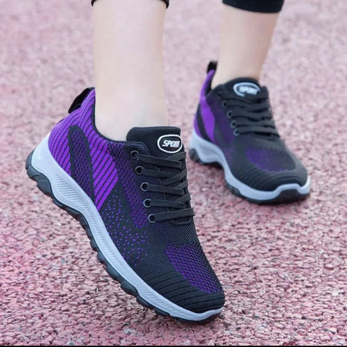 A pair of women's athletic sneakers in black and purple, shown on a person's feet on a pinkish-red textured surface. The shoes feature a black mesh upper with purple side panels, black laces, and a black and purple sole.